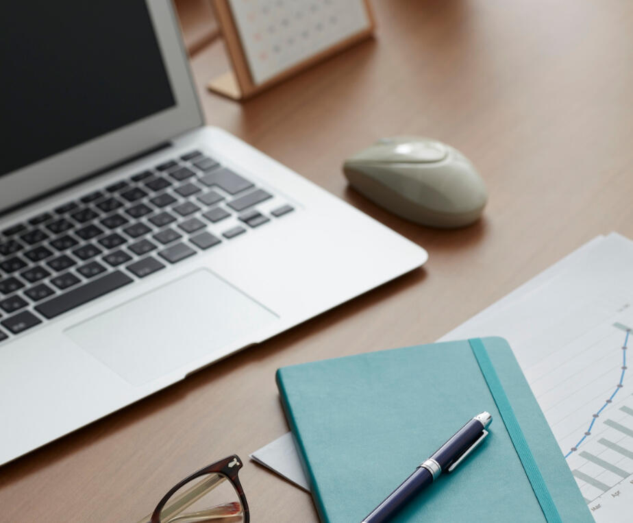 A laptop with a mouse, blue Notepad, pen and some glasses